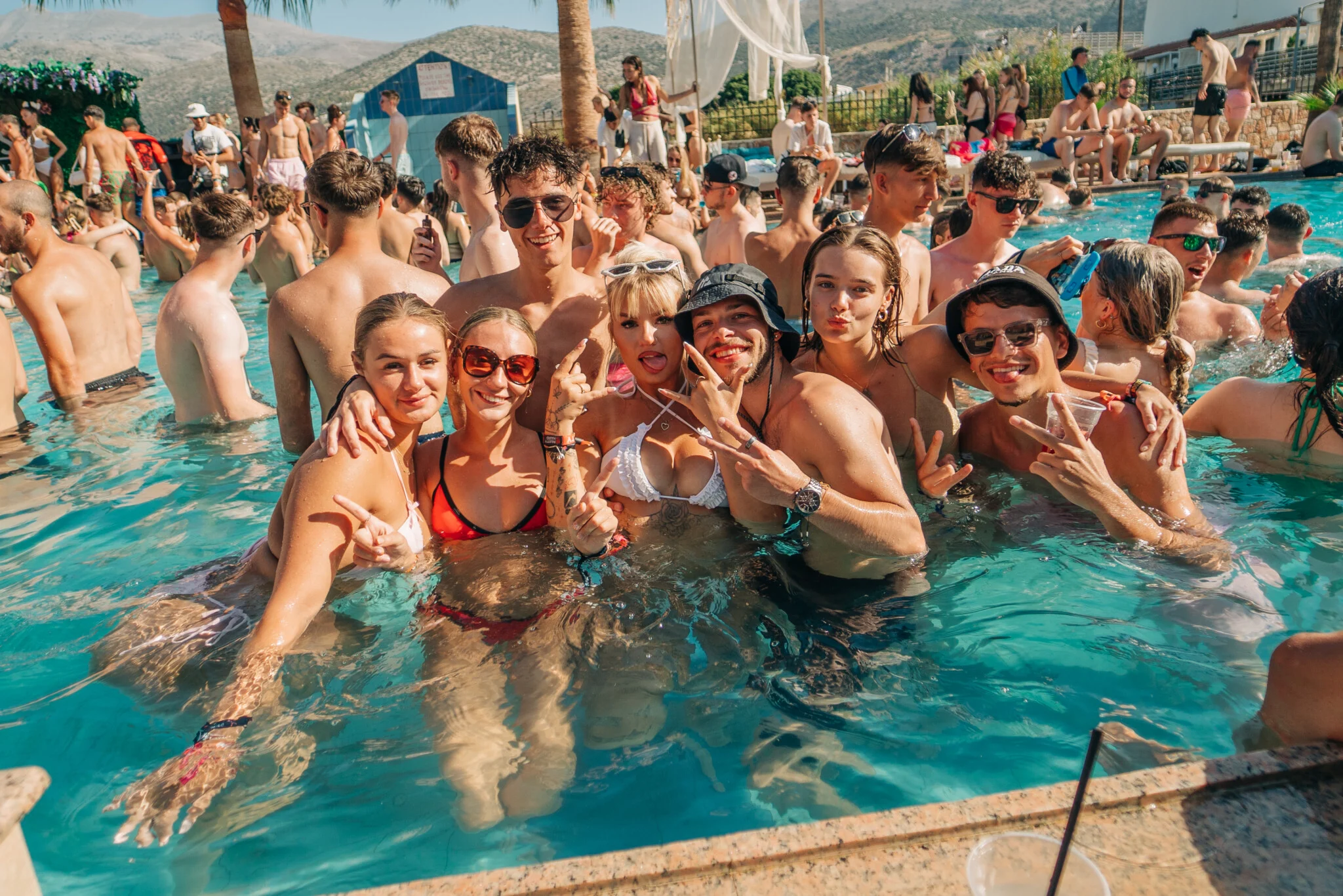 Crowds partying at the Oasis Pool Party Malia, friends posing in the water with drinks, sunshine, and full summer vibes.