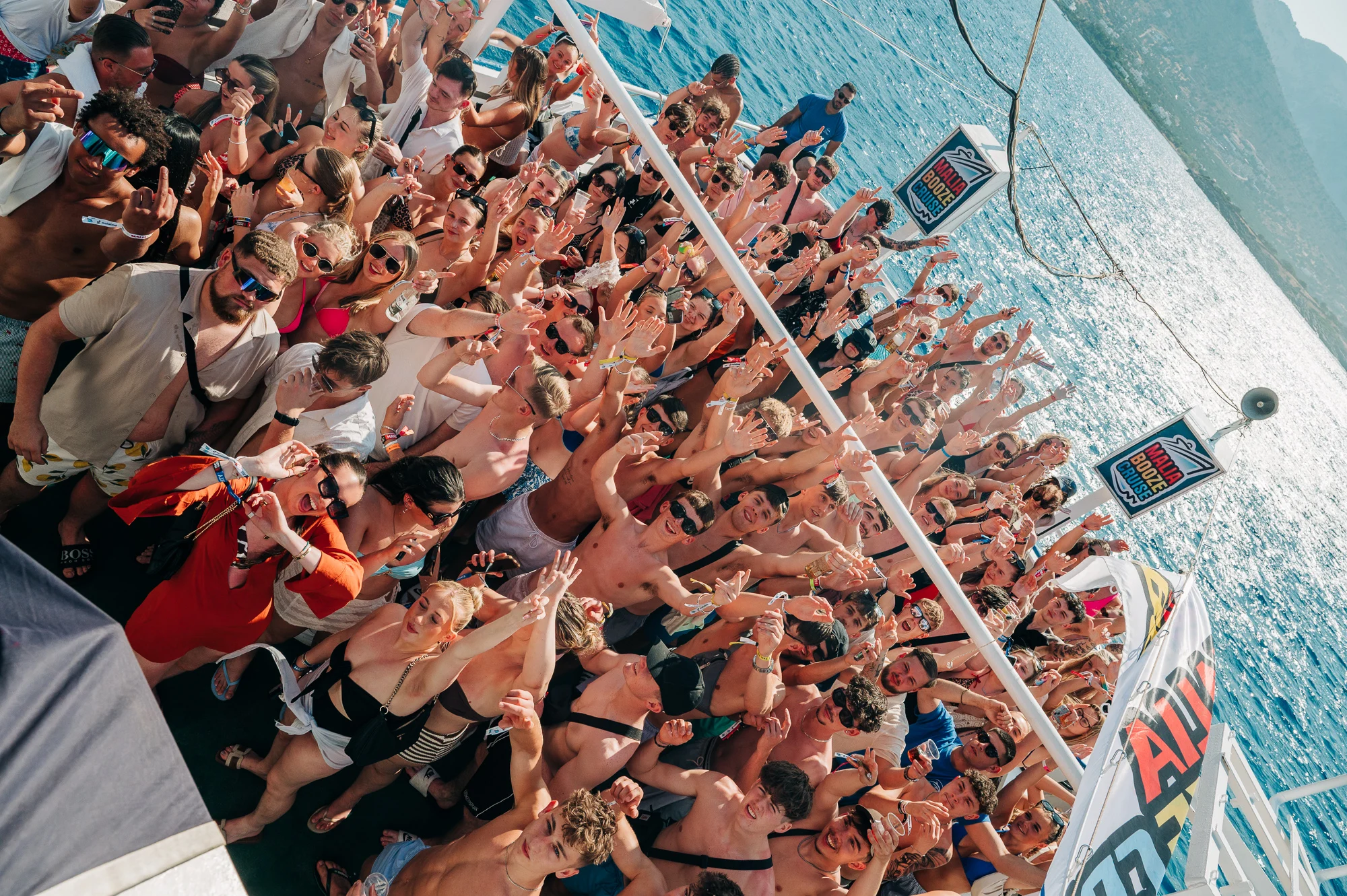 Packed top deck on the Malia Booze Cruise with hands up, drinks flowing and sea views all round—sun-soaked party vibes.