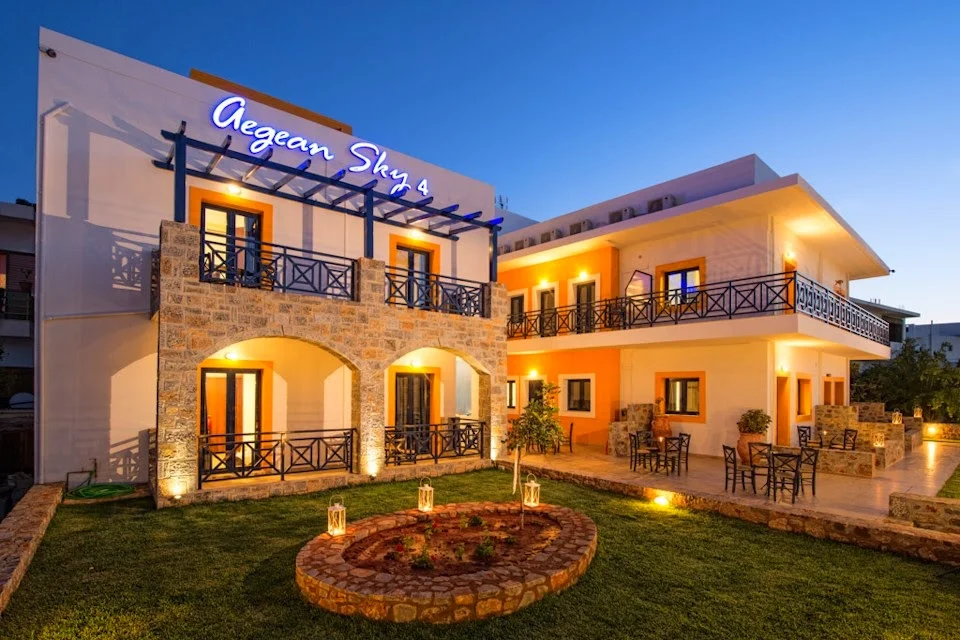 Aegean Sky Hotel & Suites, Malia—dusk scene with lit balconies, stone arches and a neat courtyard ready for your night ahead.