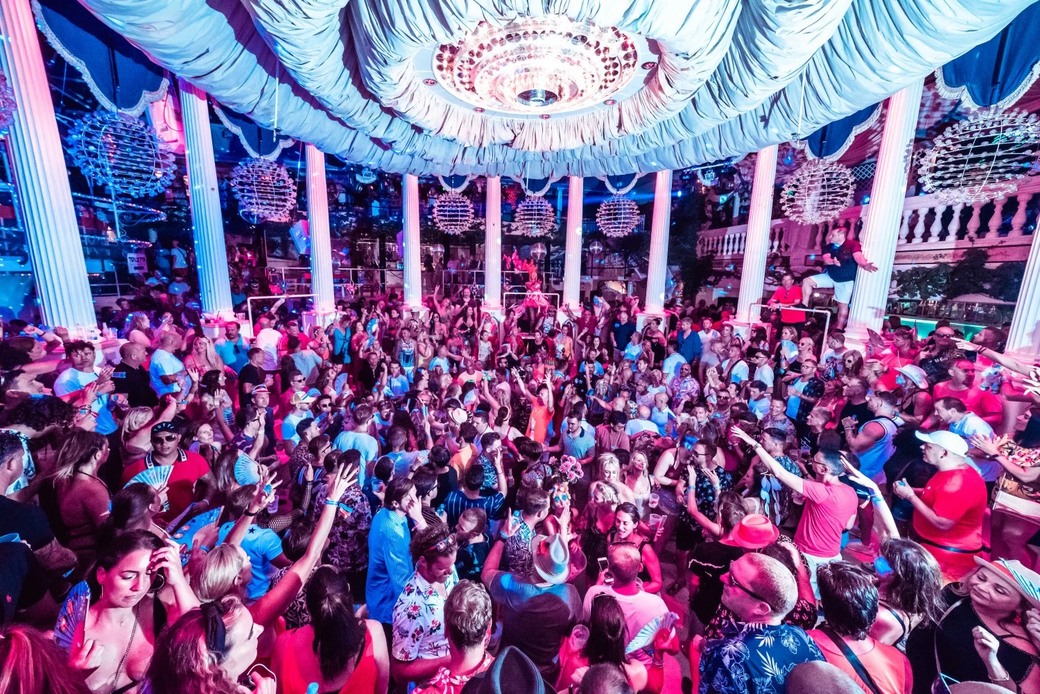 Energetic crowd dancing under chandeliers and draped ceilings at Ibiza Es Paradis club, with vibrant pink and blue lighting.