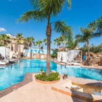 Oasis Studios Malia with palm trees, poolside beds, clear blue water and a sunny, relaxed holiday atmosphere.