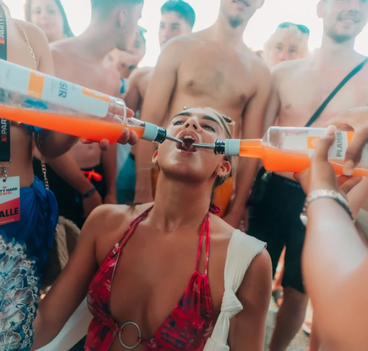 Woman at an Ayia Napa boat party drinks from two bottles at once while surrounded by a lively crowd.