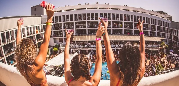 Party hard on an Ibiza clubbing holiday! Three friends raise drinks at a lively pool party with a packed club and hotel backdrop.