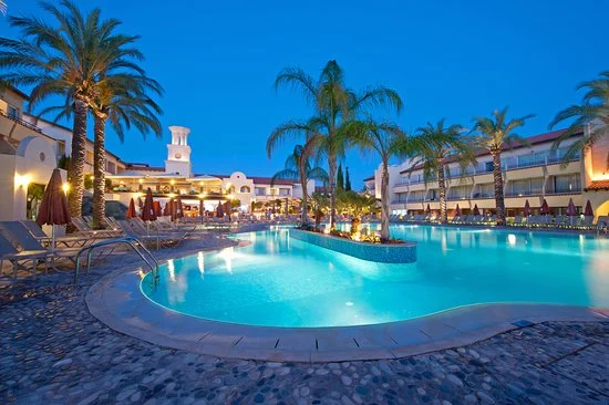 Serene pool area at a hotel in Ayia Napa, with illuminated water, palm trees, and loungers under a twilight sky.