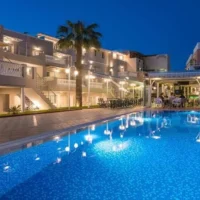 Sarpidon Apartments Malia lit up at dusk, showing the pool, balconies, palm trees and a relaxed holiday atmosphere.