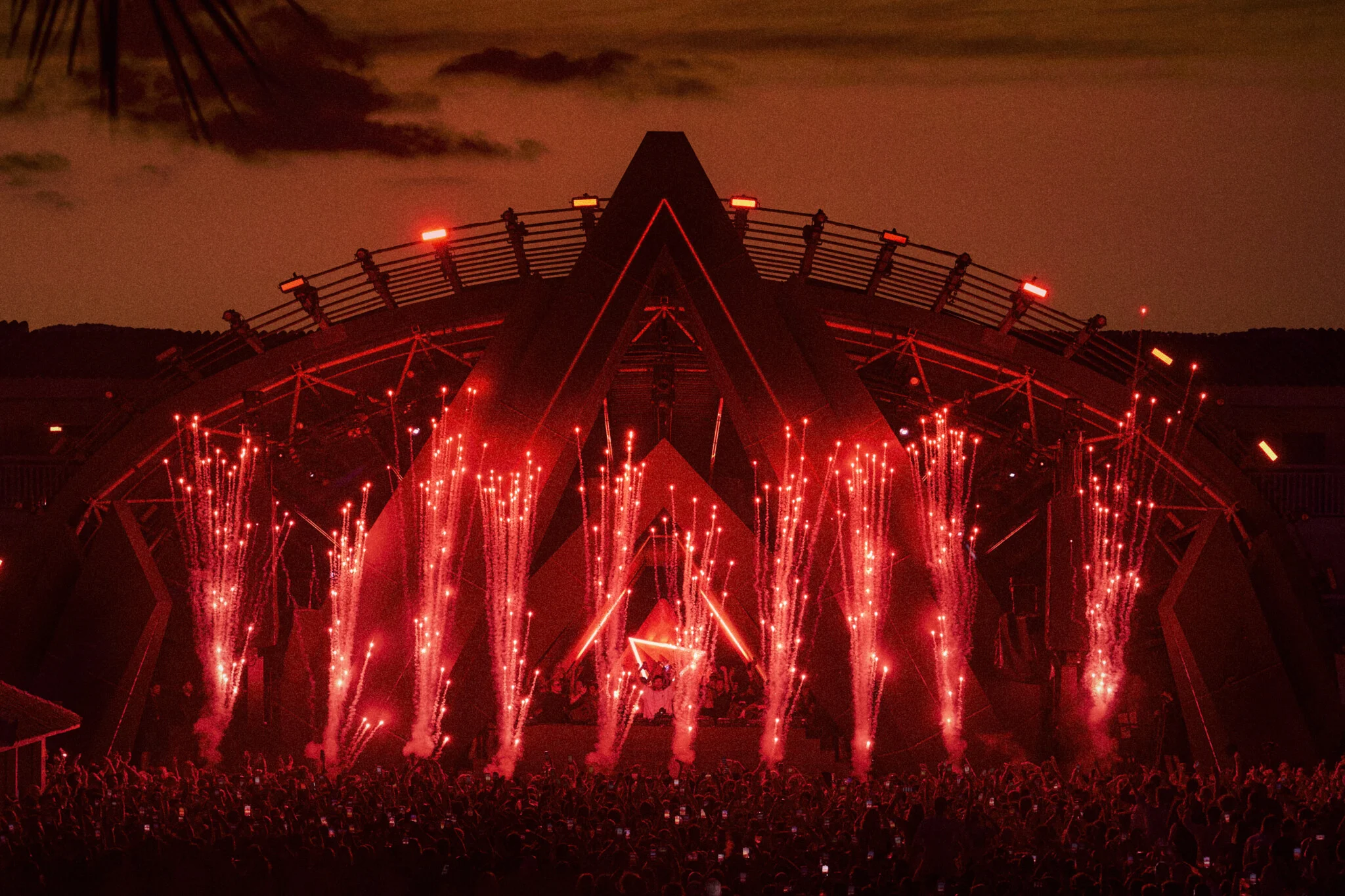 Explosive Ibiza night at Ushuaïa with fireworks, huge crowd and DJ lighting up the stage under the sunset sky.