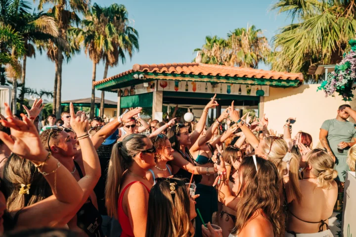 Crowds going wild under the sun at a Malia pool party, hands in the air and drinks flowing to the summer beats.