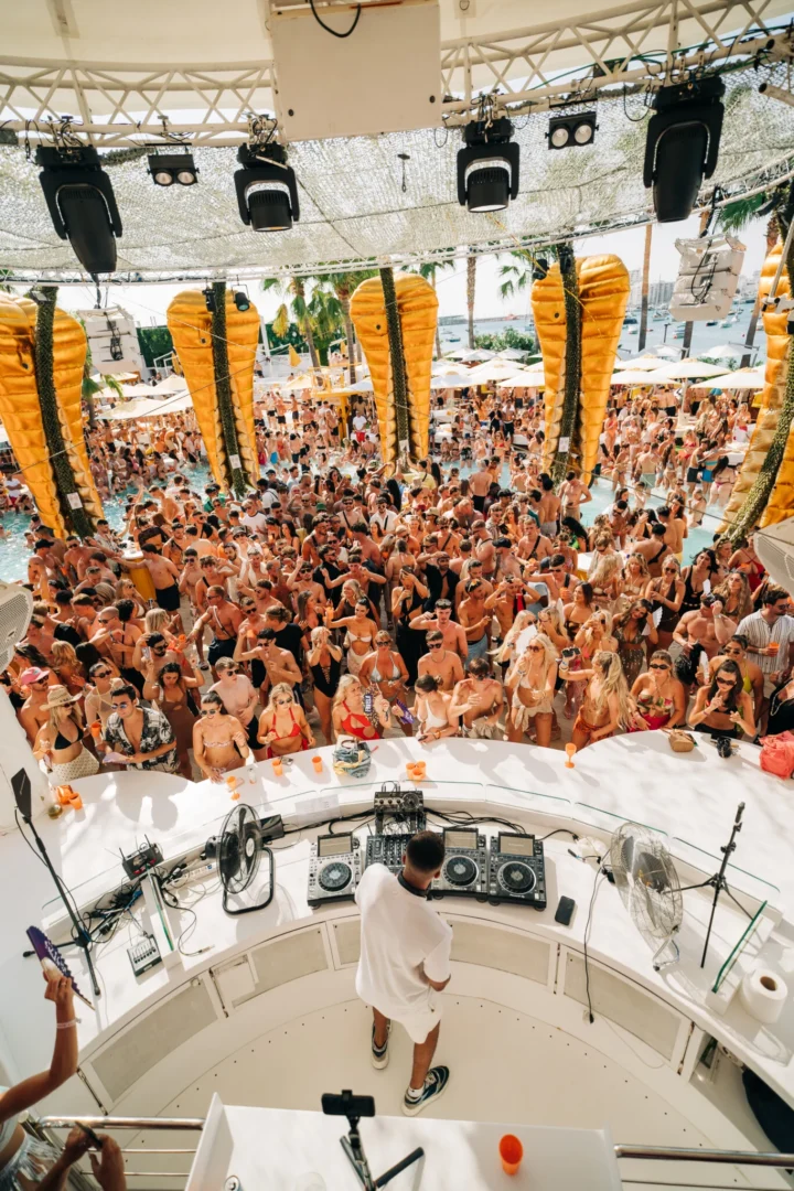 DJ performing at a packed Ibiza pool party with crowds dancing, drinks flowing, and summer vibes under the Greek sun.