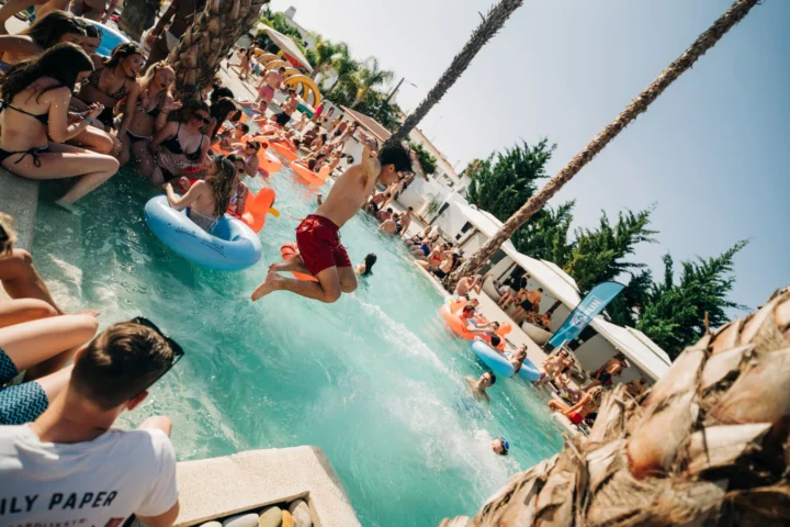 Pool party in Albufeira with people jumping in, inflatables floating and the sun shining over a buzzing crowd.