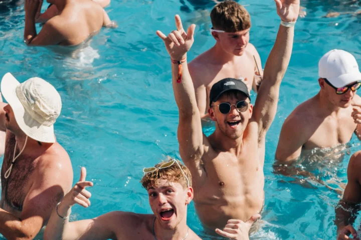 Lads buzzing in a packed Barcelona pool party, throwing up hands and cheering as the sun and music keep the energy sky-high.