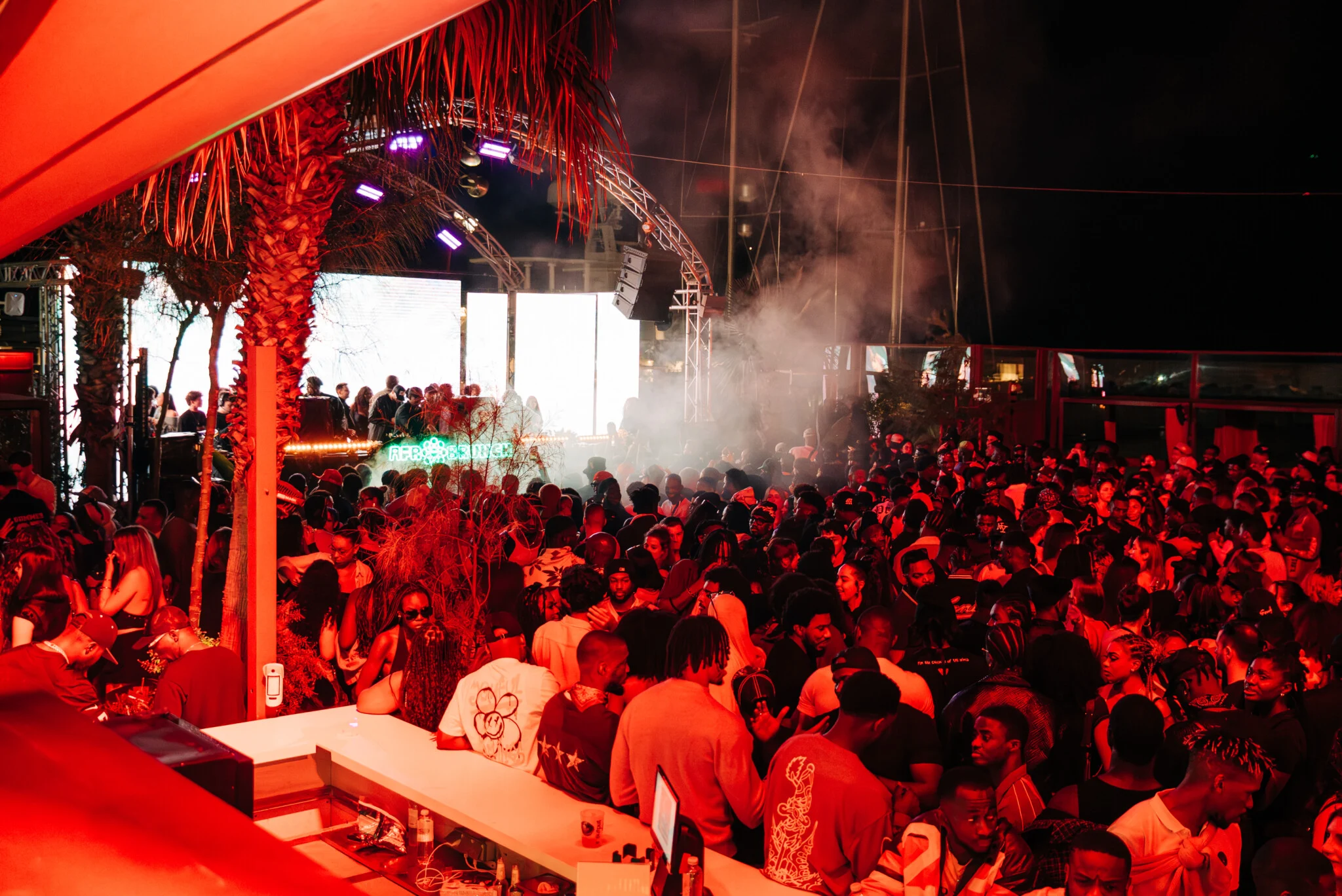 Barcelona crowd packed into a late-night club as lads and girls rave under red lights with DJs firing up the dancefloor.